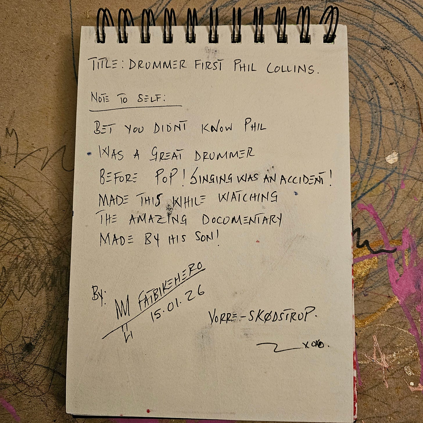 Alt Text: A handwritten note in a spiral-bound sketchbook by artist FatbikeHero. The text reads "Title: Drummer First Phil Collins. Note to self: Bet you didn't know Phil was a great drummer before pop! Singing was an accident! Made this while watching the amazing documentary made by his son!" Signed M FatbikeHero, dated 15.01.26, location Vorre-Skødstrup.