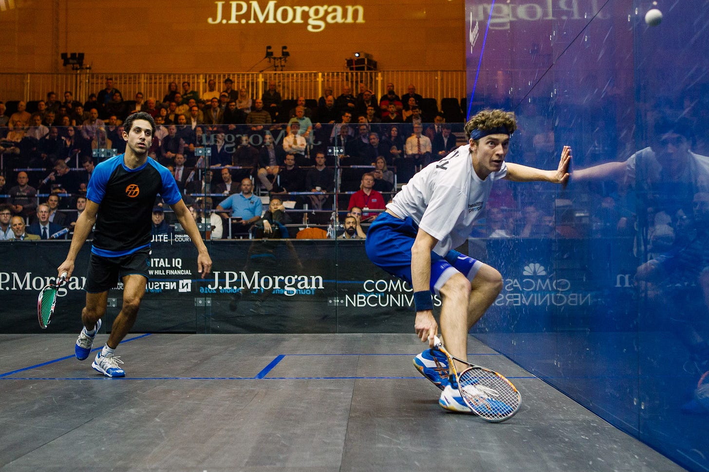 An action photo from a professional squash match inside a glass court. In the foreground, player Cameron Pilley, in a white shirt and blue shorts, lunges towards the blue side wall, his racket poised to hit the ball. In the background, his opponent watches, and a large crowd of spectators is visible behind the court. The wall has J.P. Morgan branding.