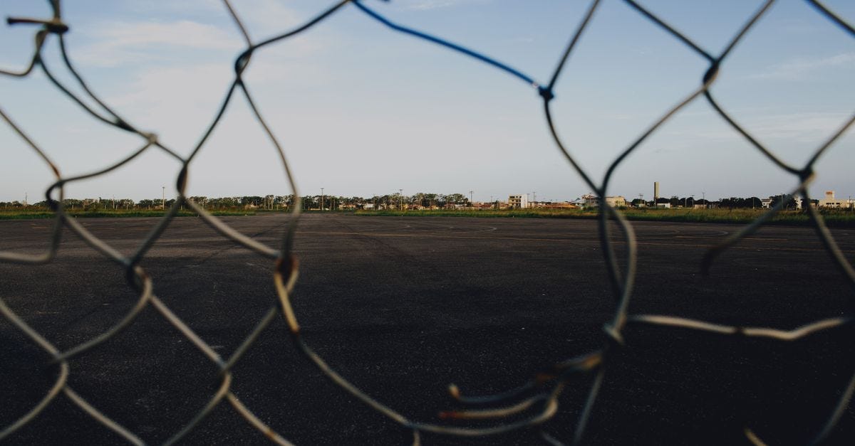 Photo: Jonathan Borba / Pexels A broken wire mesh fence framing an open asphalt area under a blue sky.