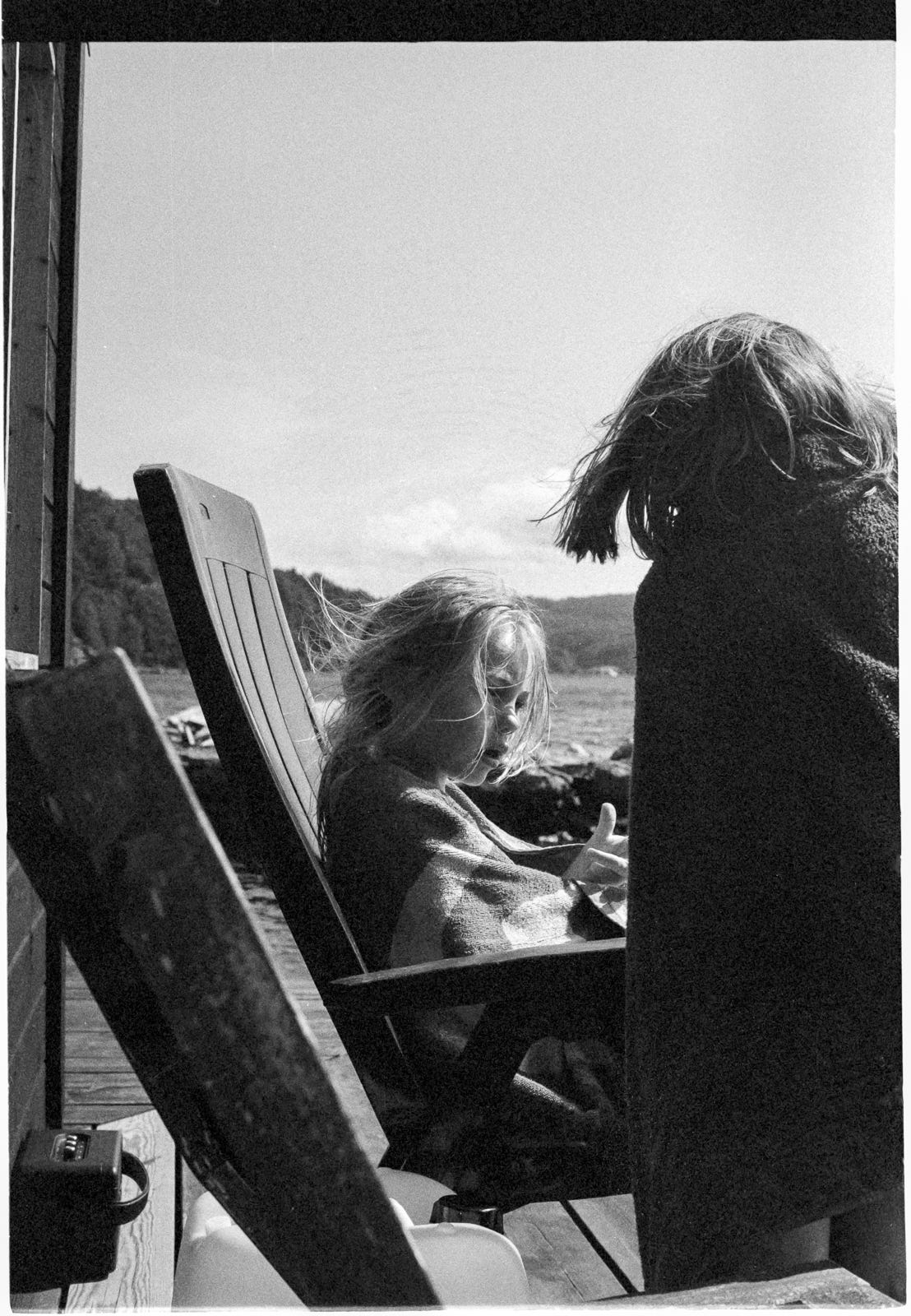 One child reclining on a chair by an ocean bay explains something to another child with serious mannerisms. Both children wear a towel. One child reclining on a chair by an ocean bay explains something to another child with serious mannerisms. Both children wear a towel.