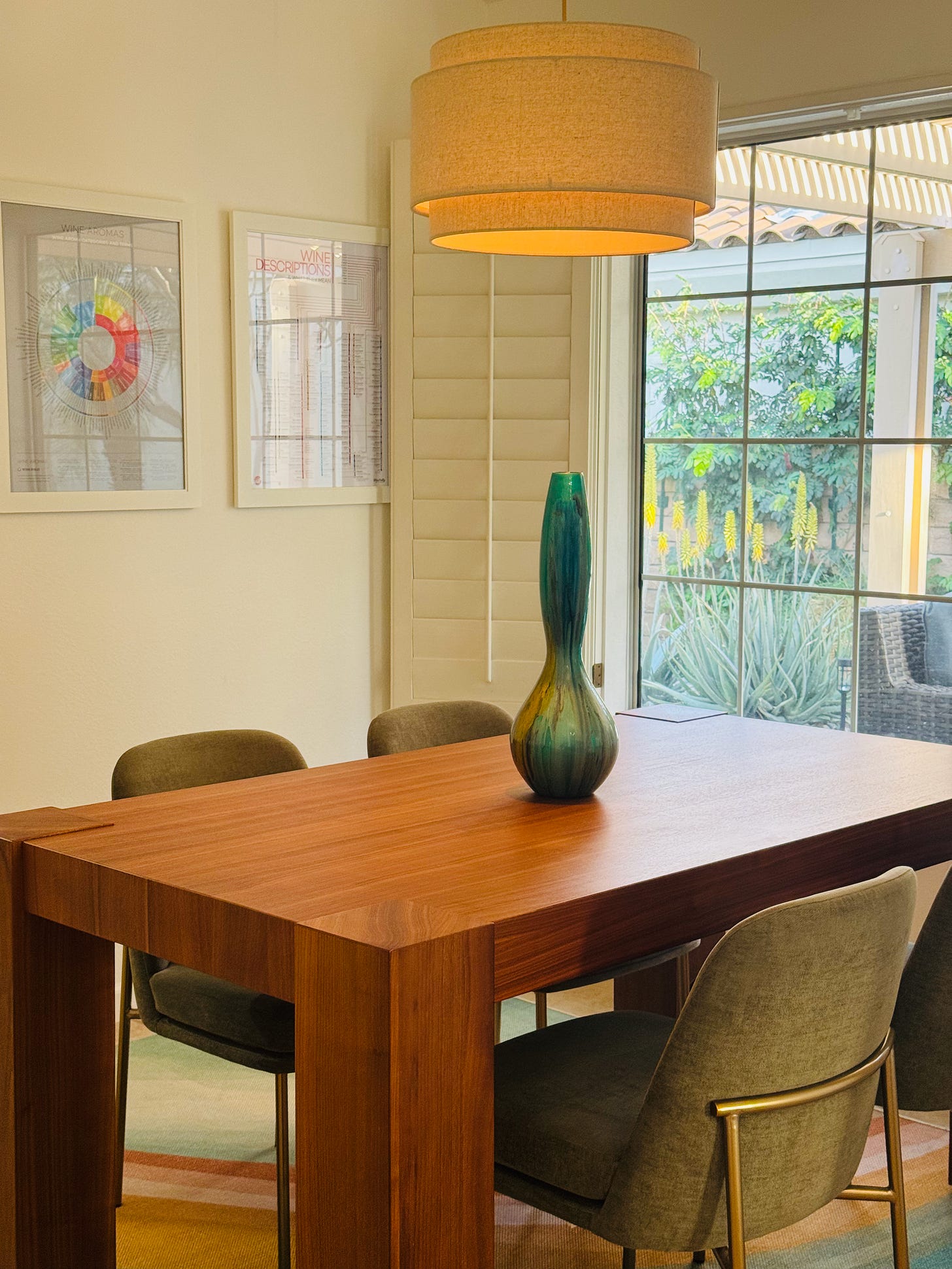 A dining room table, a modern muslin chandelier, four sage chairs, wine images on the wall, and a view to the front yard, sun shining in on the table. A dining room table, a modern muslin chandelier, four sage chairs, wine images on the wall, and a view to the front yard, sun shining in on the table.