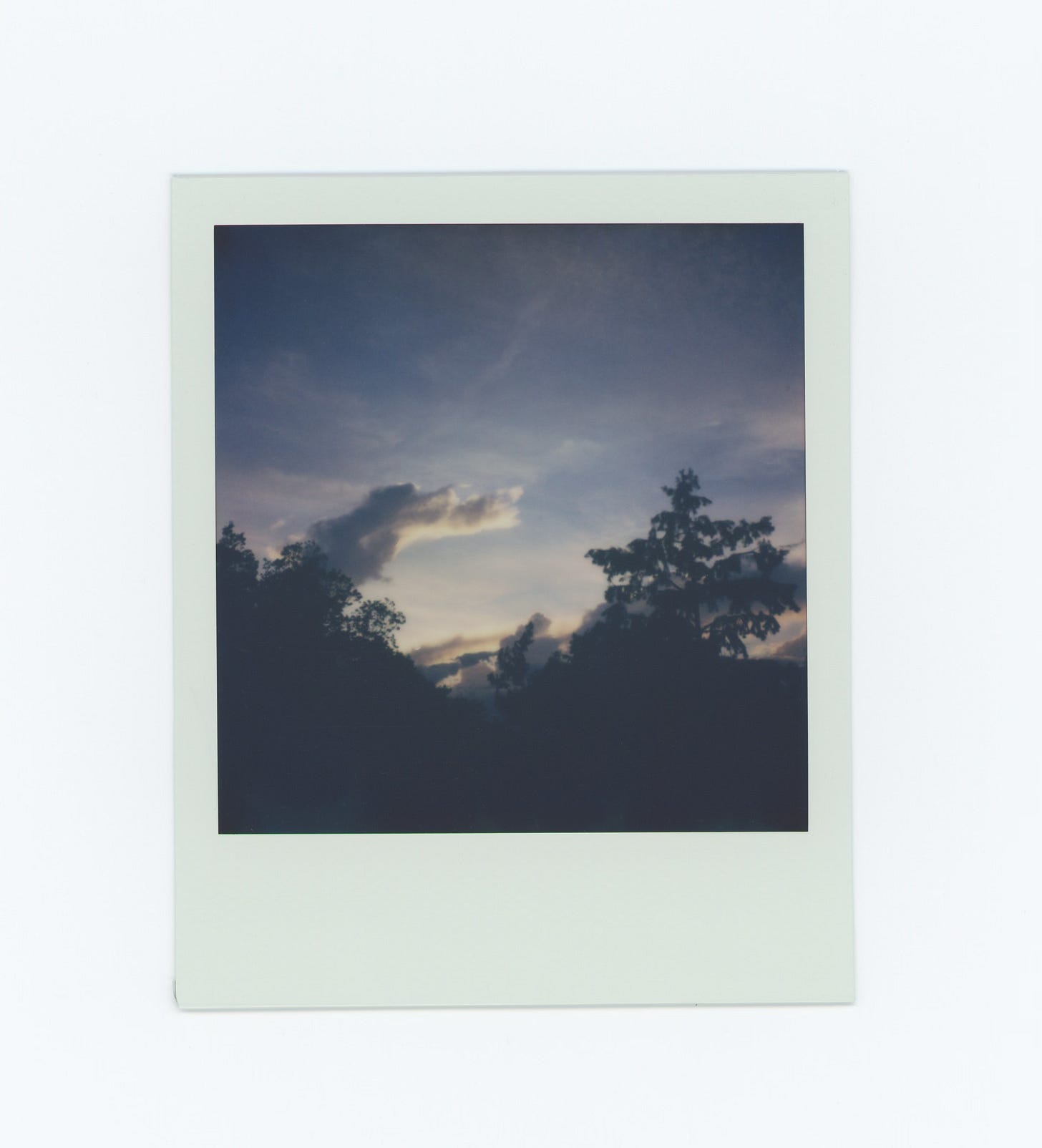 Another polaroid sunset. The sky is lighter blue in this one, and the clouds are still white around the edges. Same view, same tree line silhouette. there are some golden colored scraps of cloud right above the edge of the trees
