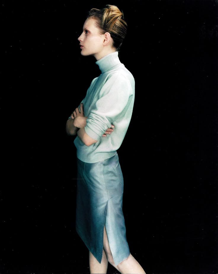 This may contain: a woman with her arms crossed standing in front of a black background wearing a white shirt and blue skirt This may contain: a woman with her arms crossed standing in front of a black background wearing a white shirt and blue skirt