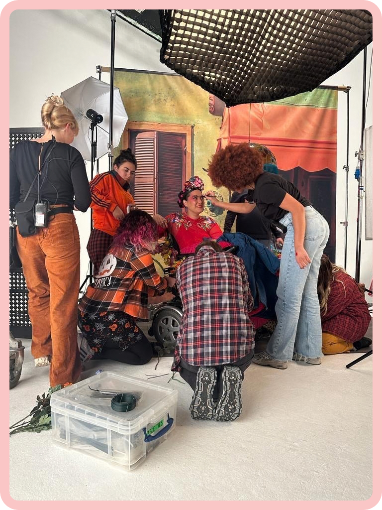 A photo of Kyla in character as Freya, who is dressed like Frida Kahlo, being surrounded by seven members of the film crew, especially hair and makeup artists. Kyla is wearing a bright red floral top, over-the-top makeup, and a floral headdress and is smiling in amusement as a woman with a red-brown afro is drawing onto her eyebrows with a pencil. Meanwhile, other people stand around Kyla, preening and adjusting different parts of her outfit. Behind and above them, large lights and boxes of film equipment suggest that we are in a film studio.