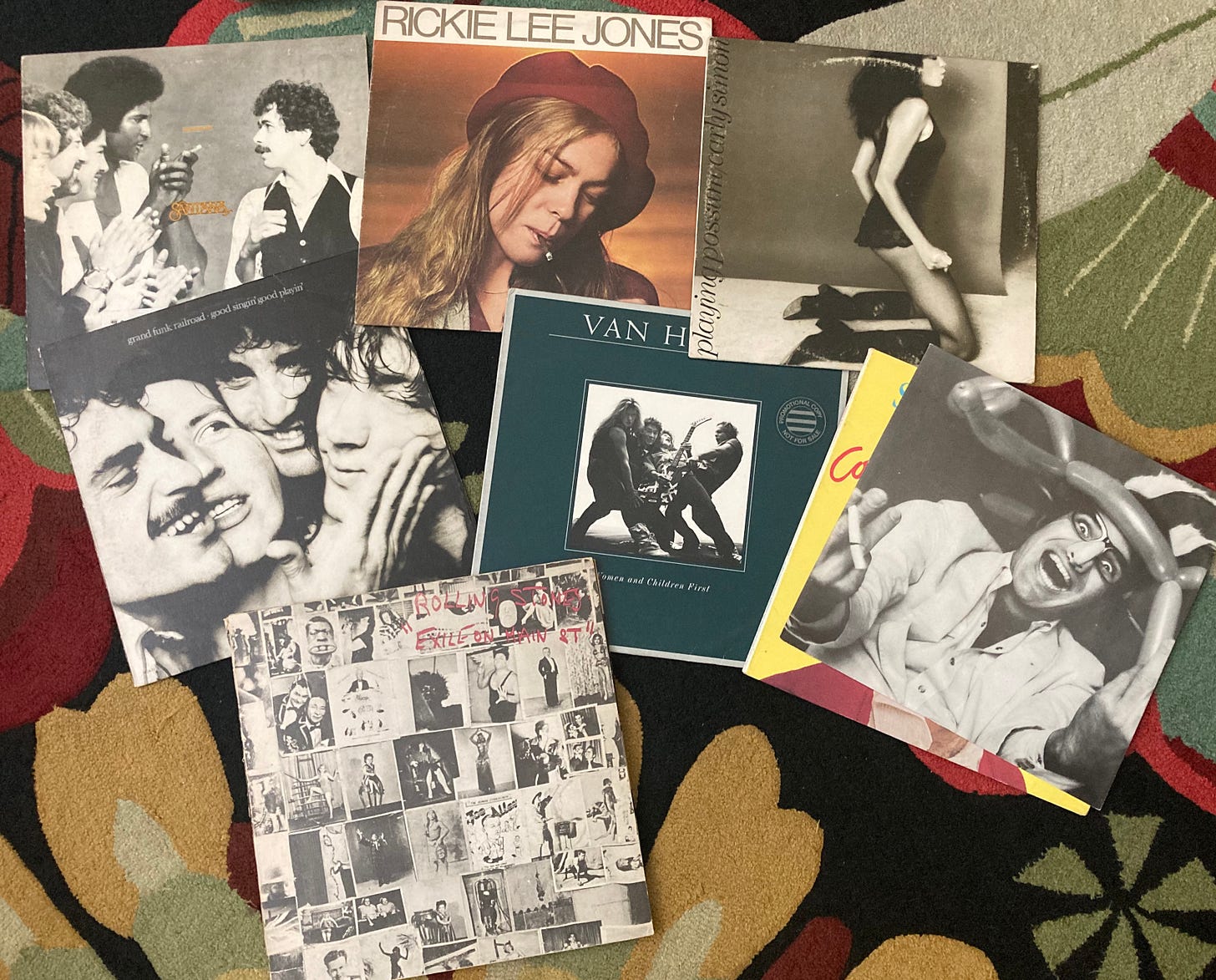A collection of seven vinyl album covers arranged on a patterned rug. Visible albums include Grand Funk Railroad’s Good Singin’ Good Playin’, Rickie Lee Jones’s self-titled debut, Carly Simon’s Playing Possum, Van Halen’s Women and Children First, Steve Martin’s Comedy Is Not Pretty!, The Rolling Stones’ Exile on Main St., and another cover featuring three men laughing in close-up. Several of these covers were photographed by Norman Seeff.