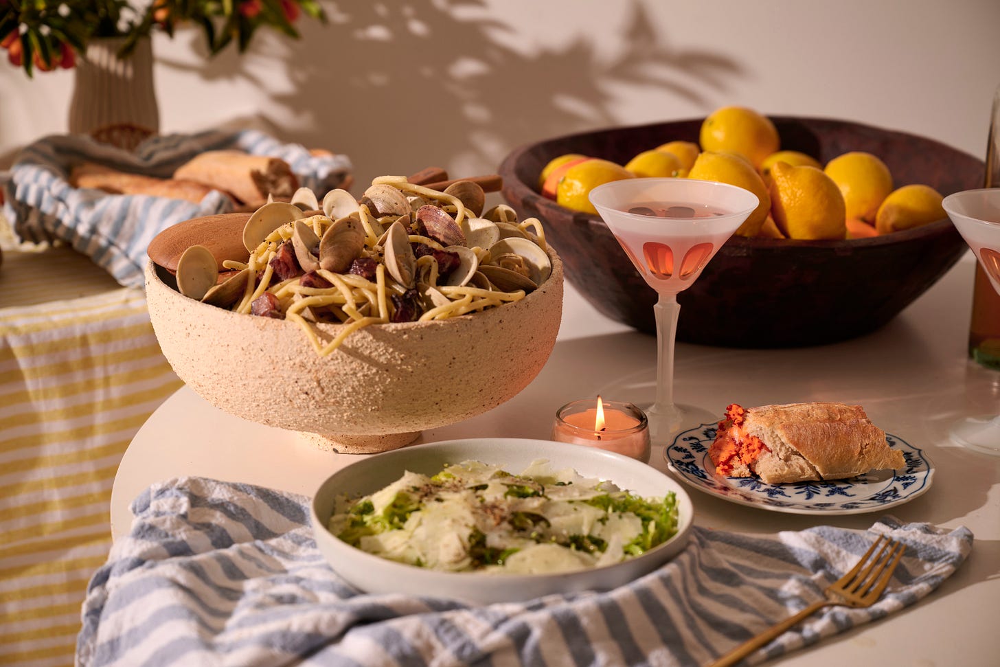 A La Dolce Vita dinner spread featuring a large bowl of spaghetti with clams and cured meat, frisée salad with shaved cheese, a slice of crusty bread with Calabrian chili butter, and a pink cocktail, set against a backdrop of lemons and fresh bread.