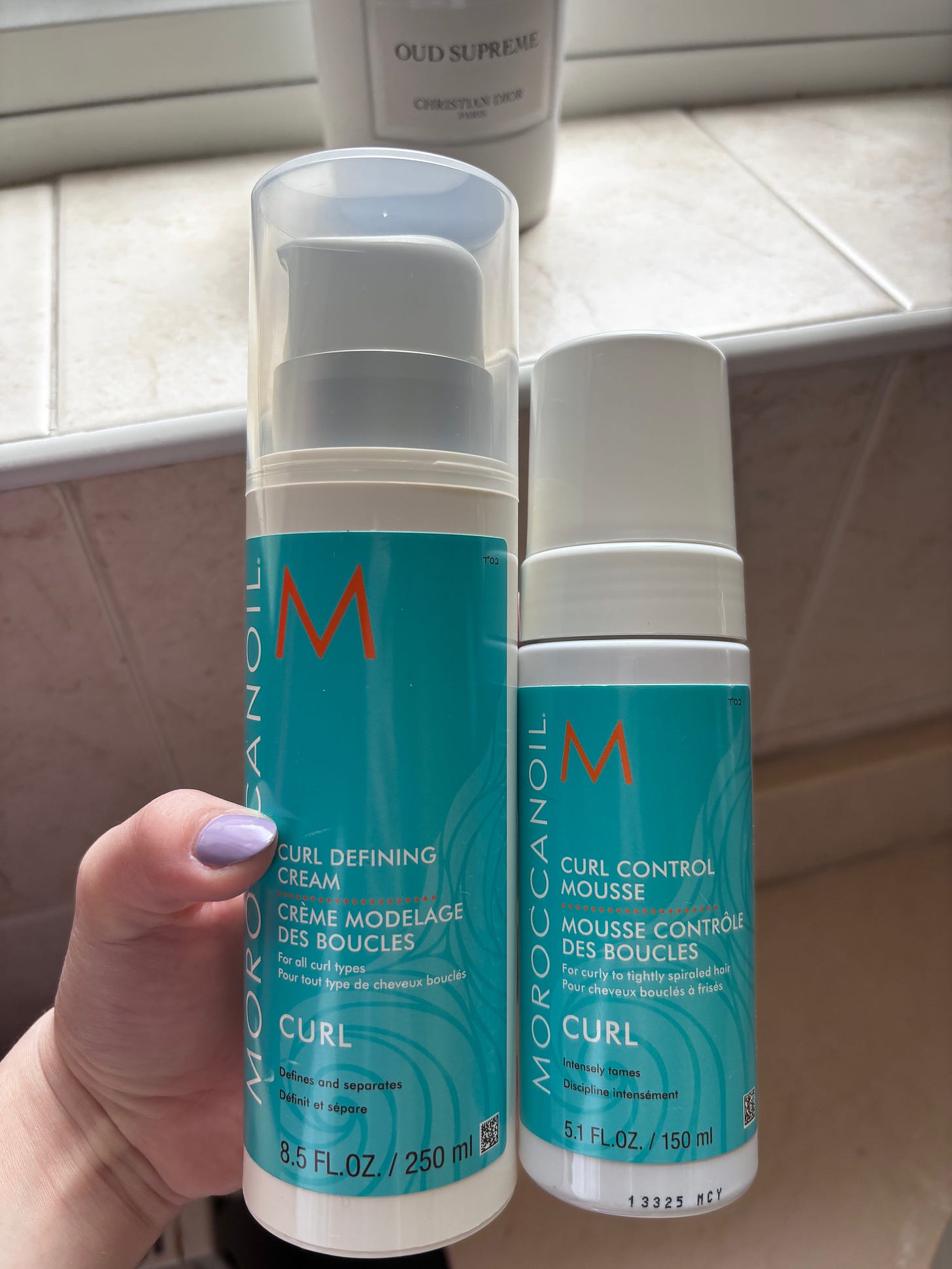 A hand holding a bottle of Moroccanoil Curl Defining Cream against a bright backdrop, highlighting the product packaging. A hand holding a bottle of Moroccanoil Curl Defining Cream against a bright backdrop, highlighting the product packaging.