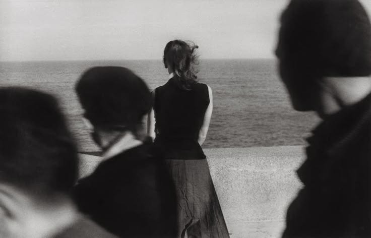 This may contain: black and white photograph of woman looking out at the ocean with people standing around her