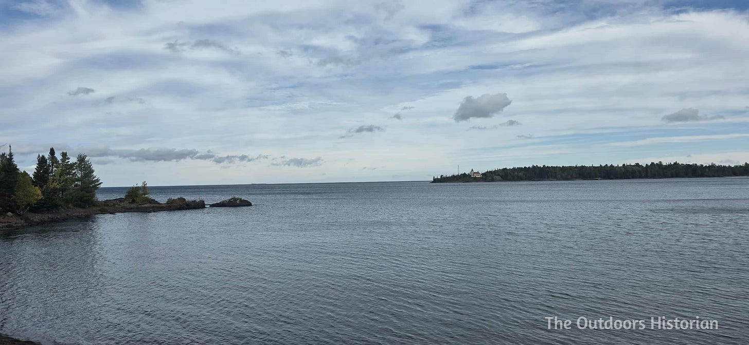 A view of a Lake Superior with a closer rocky point on the right hand side and a rocky point that is further away on the left hand side. The latter has a lighthouse on it. The sky is blue with many feathery white clouds in front of it.