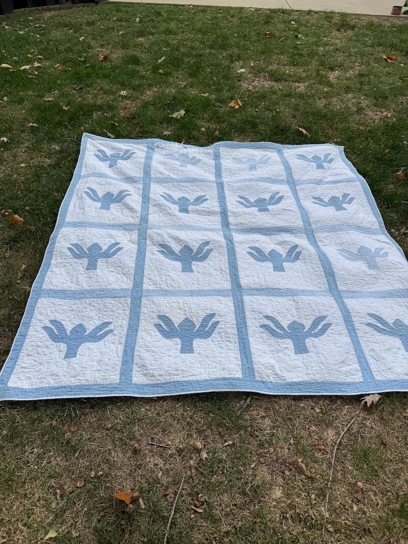 May include: A handmade quilt with a repeating pattern of light blue stylized bird shapes on a white background. The quilt is bordered and sectioned with light blue fabric. The quilt is displayed on a grassy surface. May include: A handmade quilt with a repeating pattern of light blue stylized bird shapes on a white background. The quilt is bordered and sectioned with light blue fabric. The quilt is displayed on a grassy surface.
