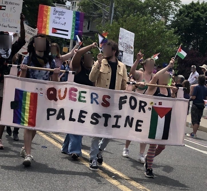 A group of people marching down a street, carrying a homemade sign that says QUEERS FOR PALESTINE, with a rainbow flag and Palestinian flag sewn on either side. People in the crowd are holding up both rainbow and Palestinian flags  