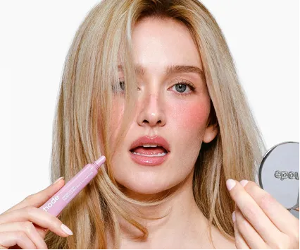 Sarah Pidgeon applying Rhode Peptide Lip Tint while looking into a compact mirror, showcasing a sheer, luminous makeup look.