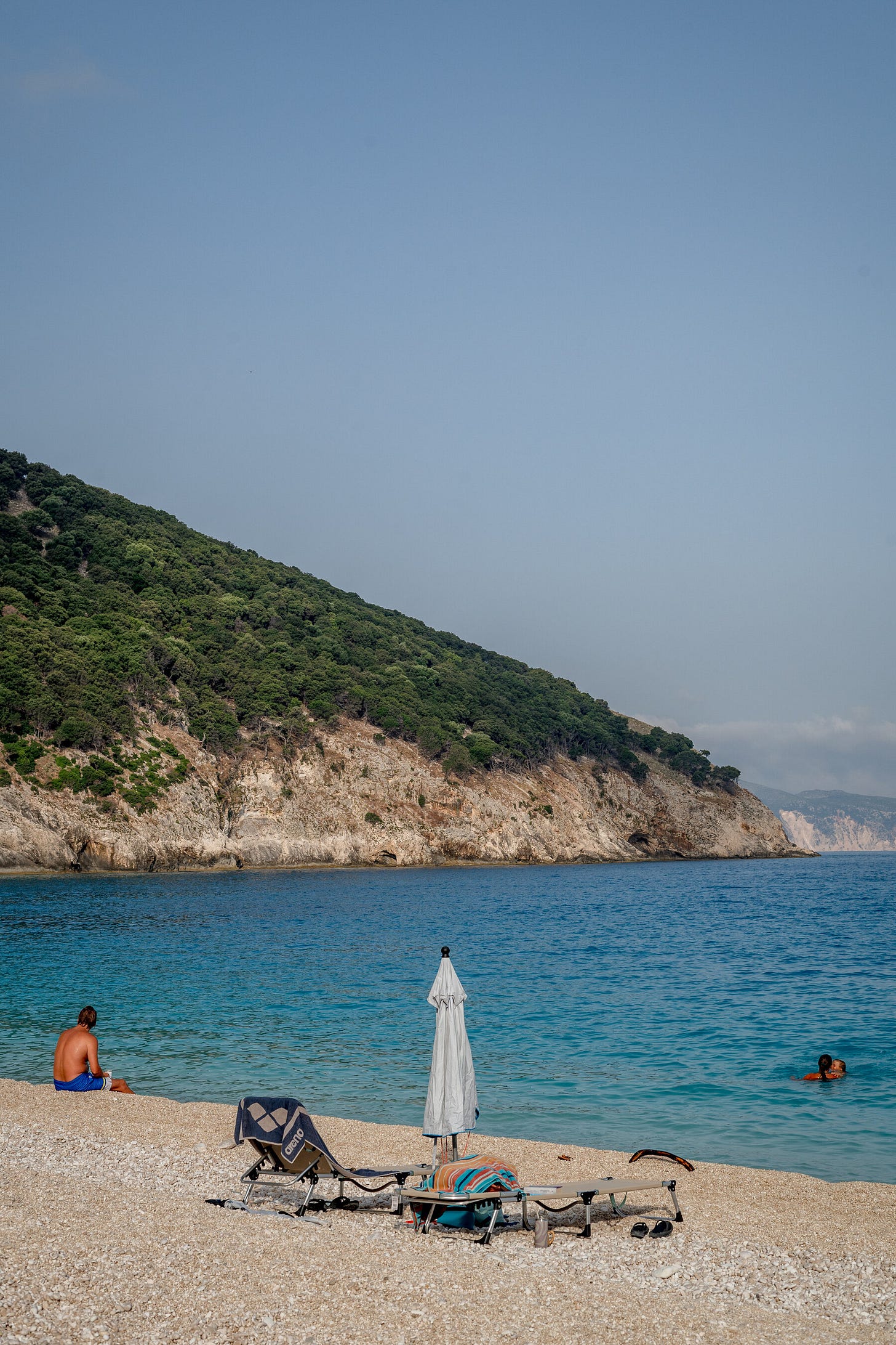 A person sits on a pebbly shore at a beach surrounded by sheer cliffs.