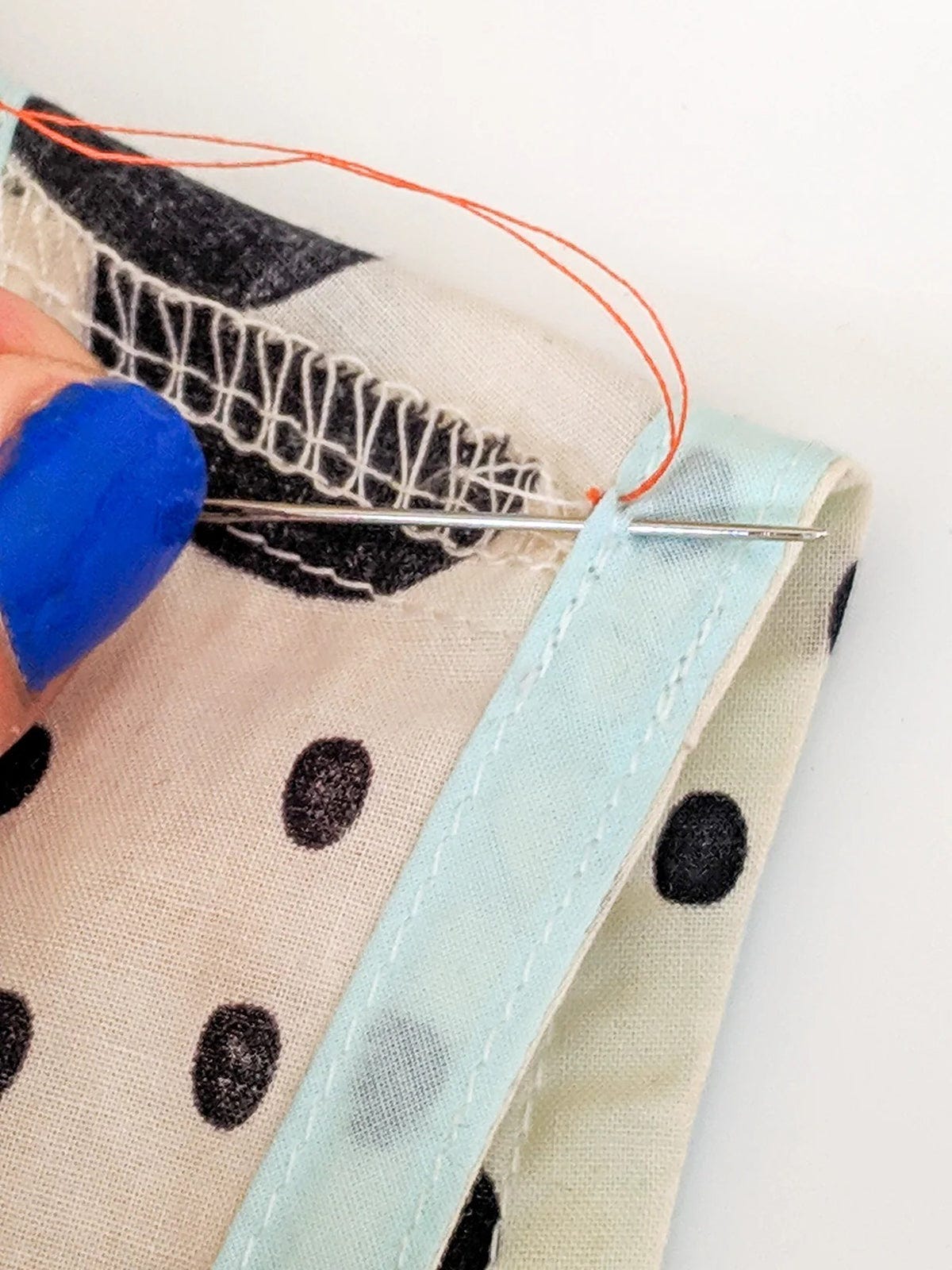 Needle pulling orange thread through fabric near armhole seam with serged edge and light blue binding.