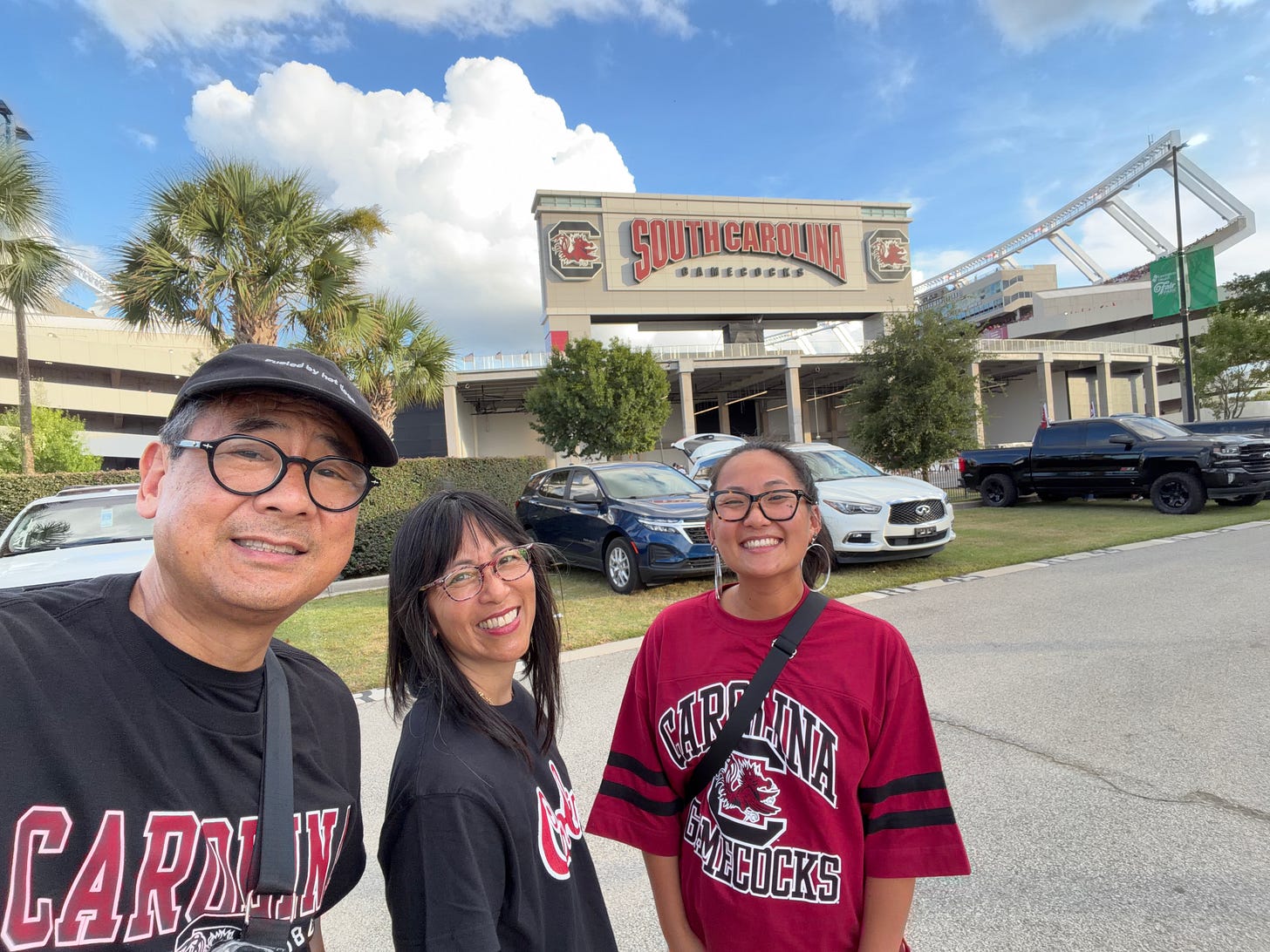 Selfie of three smiling people standing outside Williams-Brice Stadium on a sunny day, all wearing South Carolina Gamecocks shirts, with the stadium's team sign visible behind them. Selfie of three smiling people standing outside Williams-Brice Stadium on a sunny day, all wearing South Carolina Gamecocks shirts, with the stadium's team sign visible behind them.