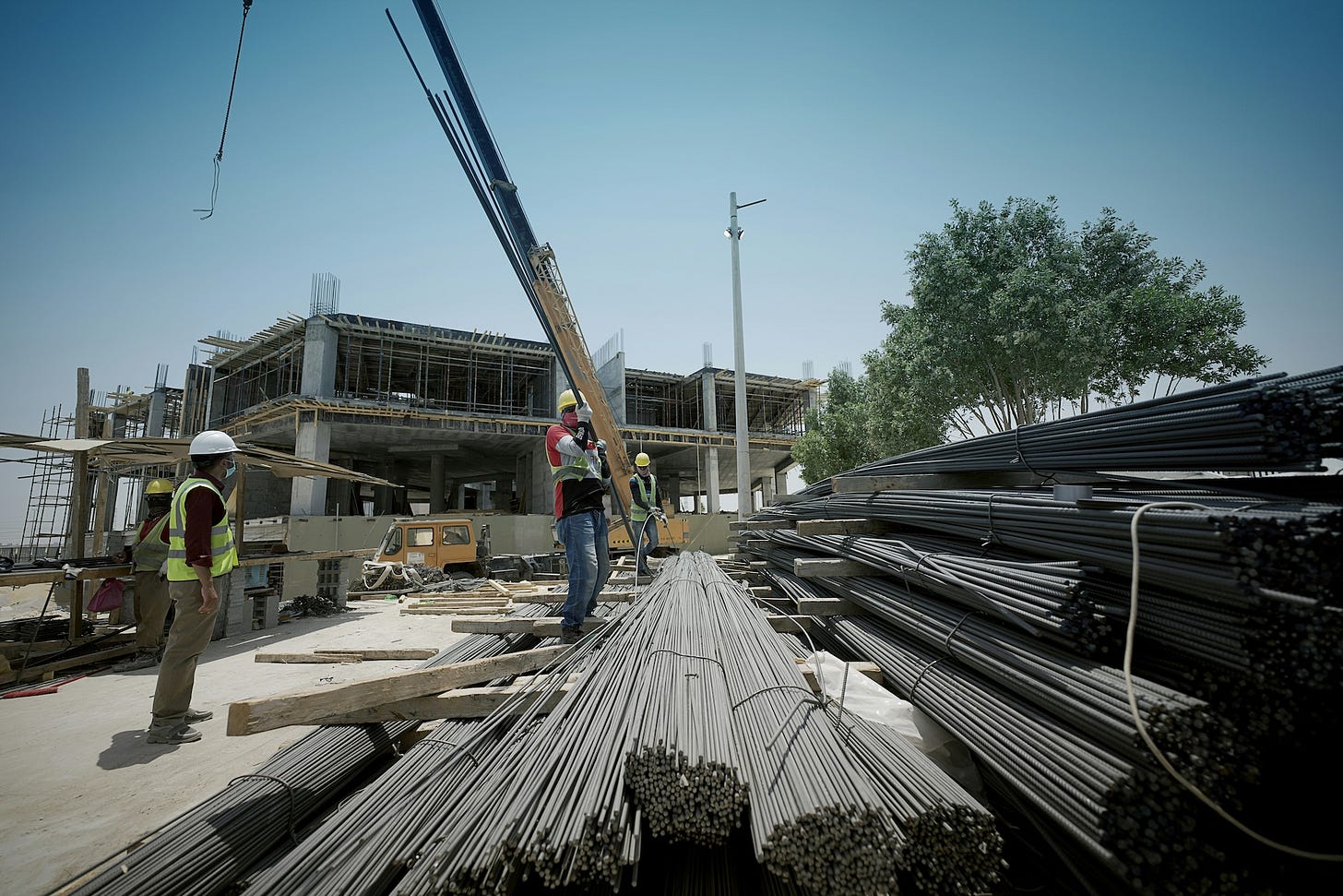 Construction site with workers, mobile crane, stacked rebar, and a multi-level building under construction in a residential area.