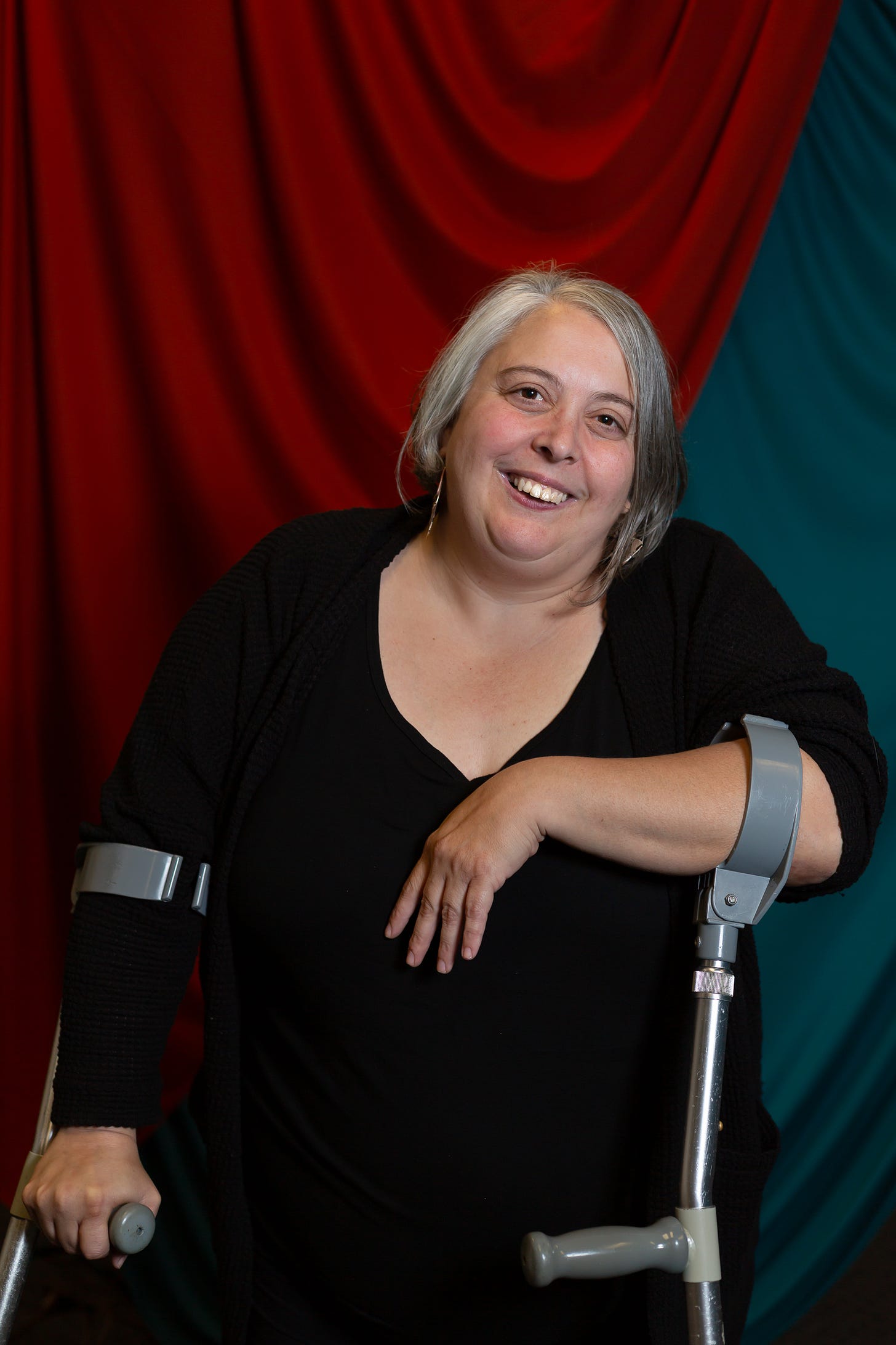 Portrait of a white skinned woman with a short gray bob. She has brown eyes, silver hoops, and is smiling at the cameral. She is wearing a long sleeved black shirt and resting on forearm crutches.
