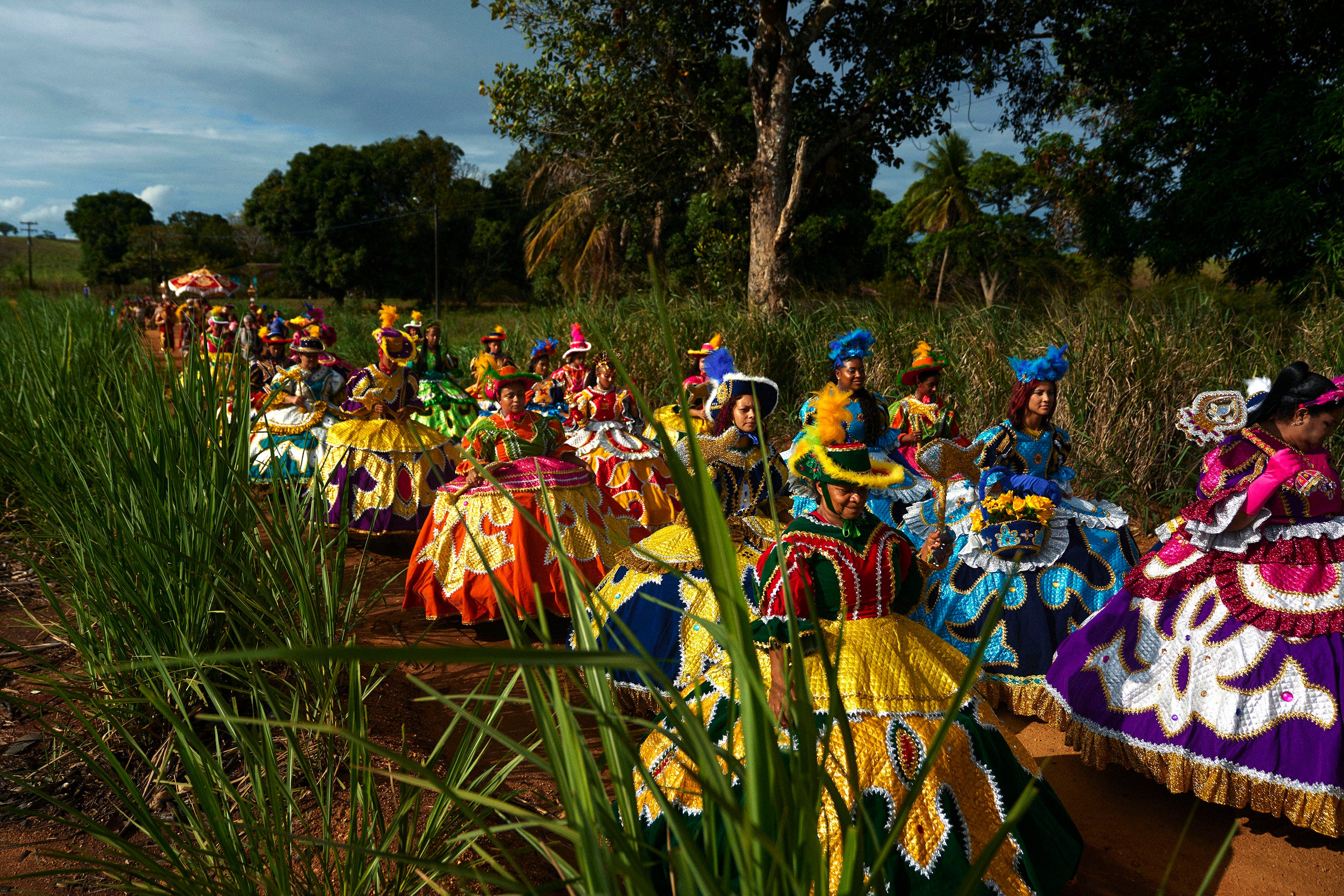 A group of people wearing brightly colored, elaborate traditional costumes walk in a line along a dirt path through tall grass and trees on a sunny day.