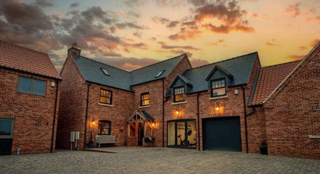 A large brick house with black windows and a grey roof, lights hanging on it, a grey driveway, and a sunset sky above it