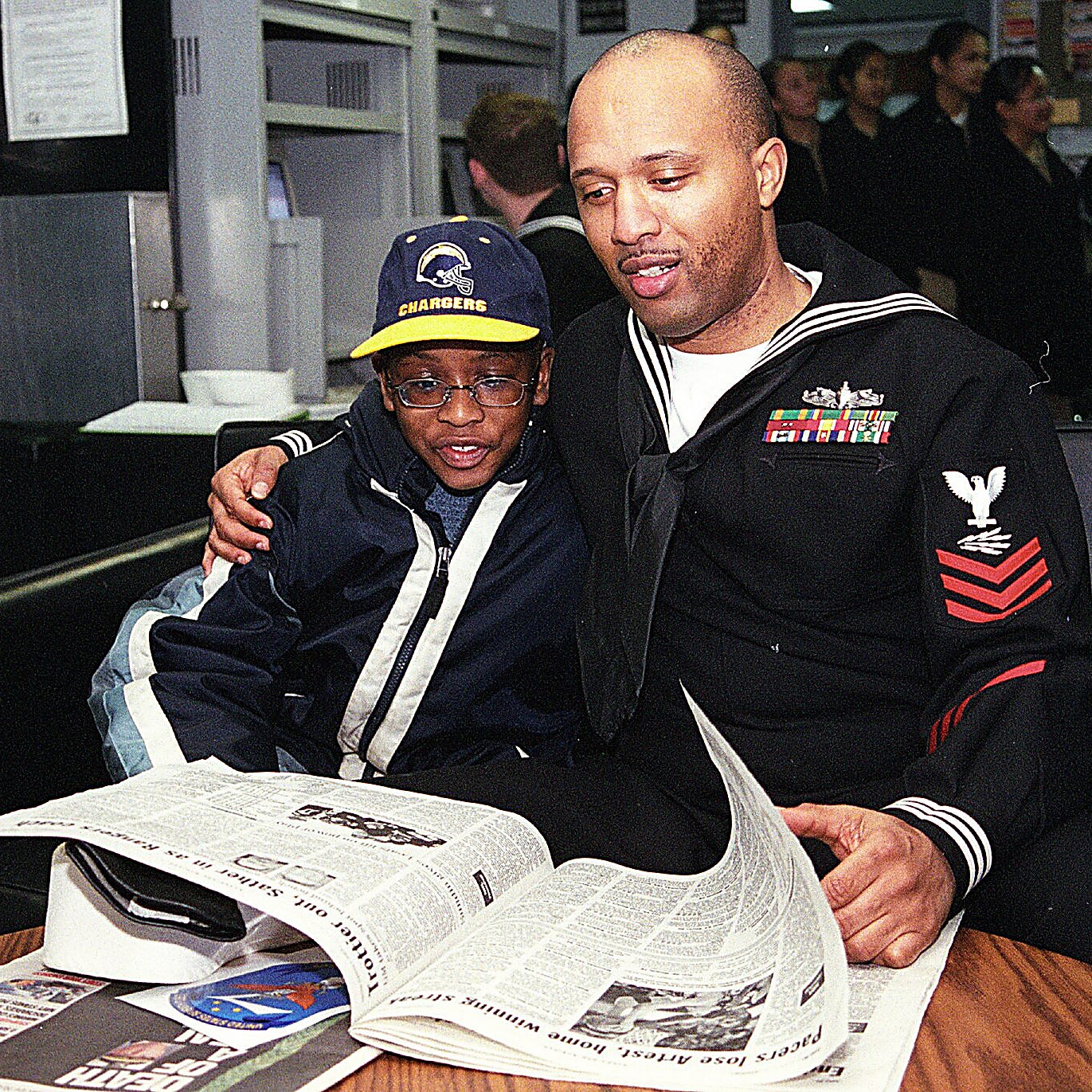 A Navy petty officer in dress blues sits with his arm around a young boy wearing glasses and a San Diego Chargers cap; an open newspaper is spread on the table in front of them. A Navy petty officer in dress blues sits with his arm around a young boy wearing glasses and a San Diego Chargers cap; an open newspaper is spread on the table in front of them.