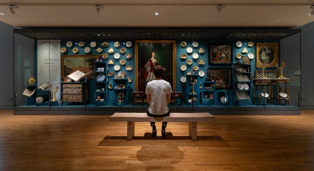 A person sat on a wooden bench looking at an art collection with plates and paintings with a blue backdrop A person sat on a wooden bench looking at an art collection with plates and paintings with a blue backdrop