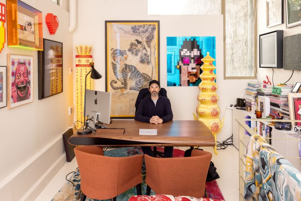 A man sits at a desk in a colorful office-like gallery space surrounded by contemporary artworks, books, a tiger image, pixelated portraiture and sculptural objects.