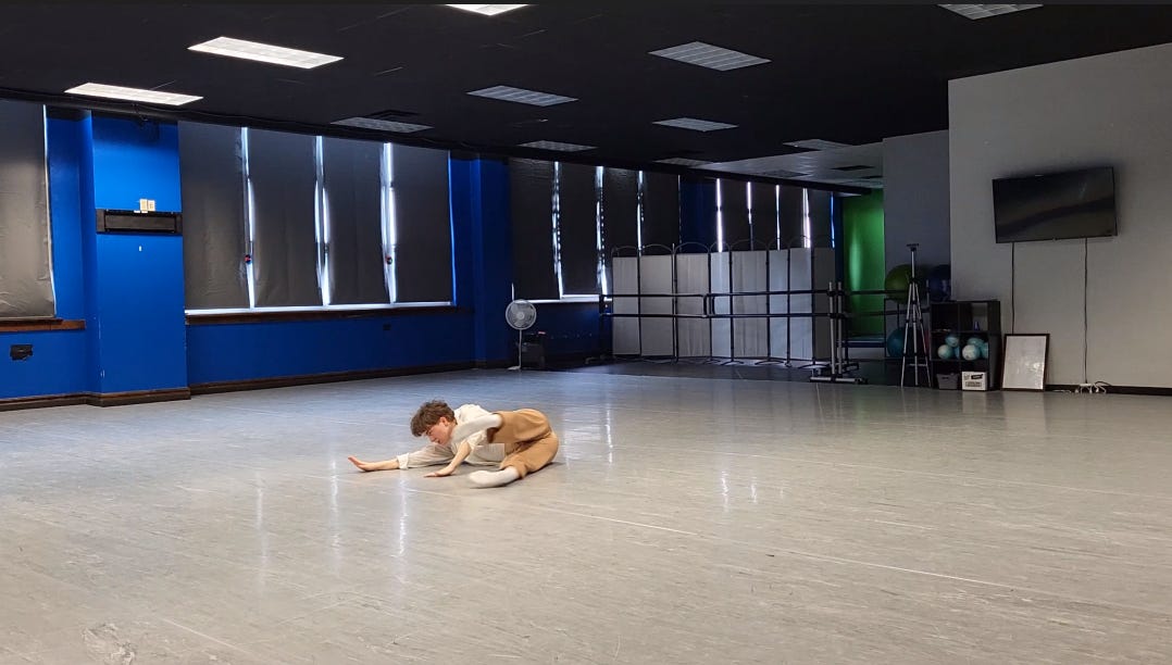 Gillian, a nonbinary white person with brown hair and pale skin, lays on their right side on the floor, arms reaching along the ground as their gaze follows. They extend their legs at a ninety degree angle with their feet flexed. They wear a white button down with tan pants and white socks.