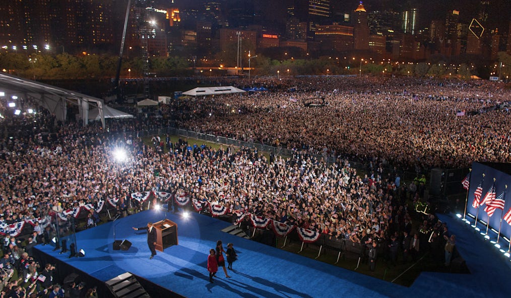 A huge crowd stretches across a large park, lit up by bright lights. At the lower left-hand corner, President Barack Obama waves at the crowd, while his wife and daughters walk off a large stage carpeted in blue. A huge crowd stretches across a large park, lit up by bright lights. At the lower left-hand corner, President Barack Obama waves at the crowd, while his wife and daughters walk off a large stage carpeted in blue.