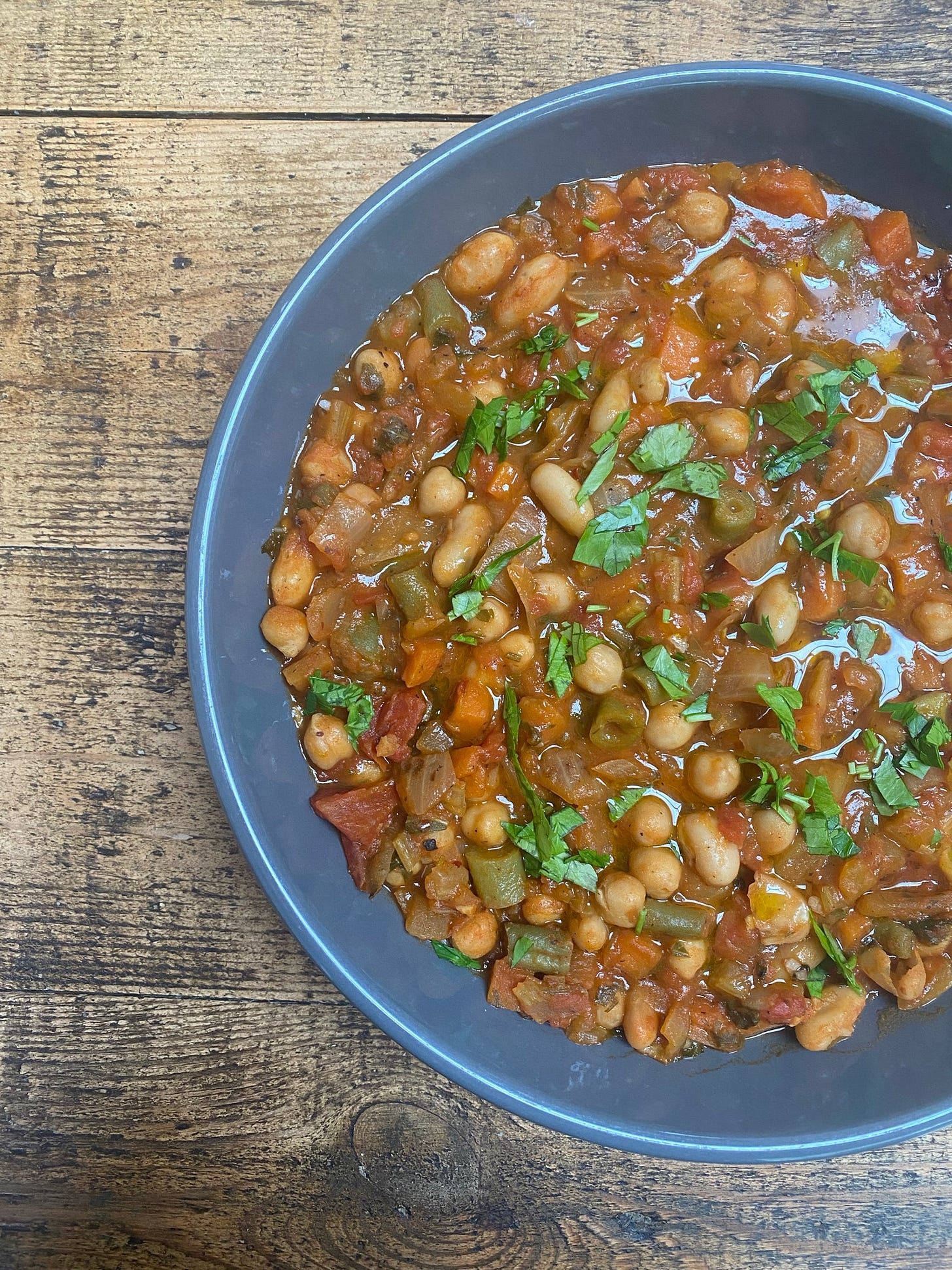 A grey bowl is on a wooden table. In the bowl there are beans and vegetables in a tomato sauce. The dish is scattered with fresh herbs, which are green.