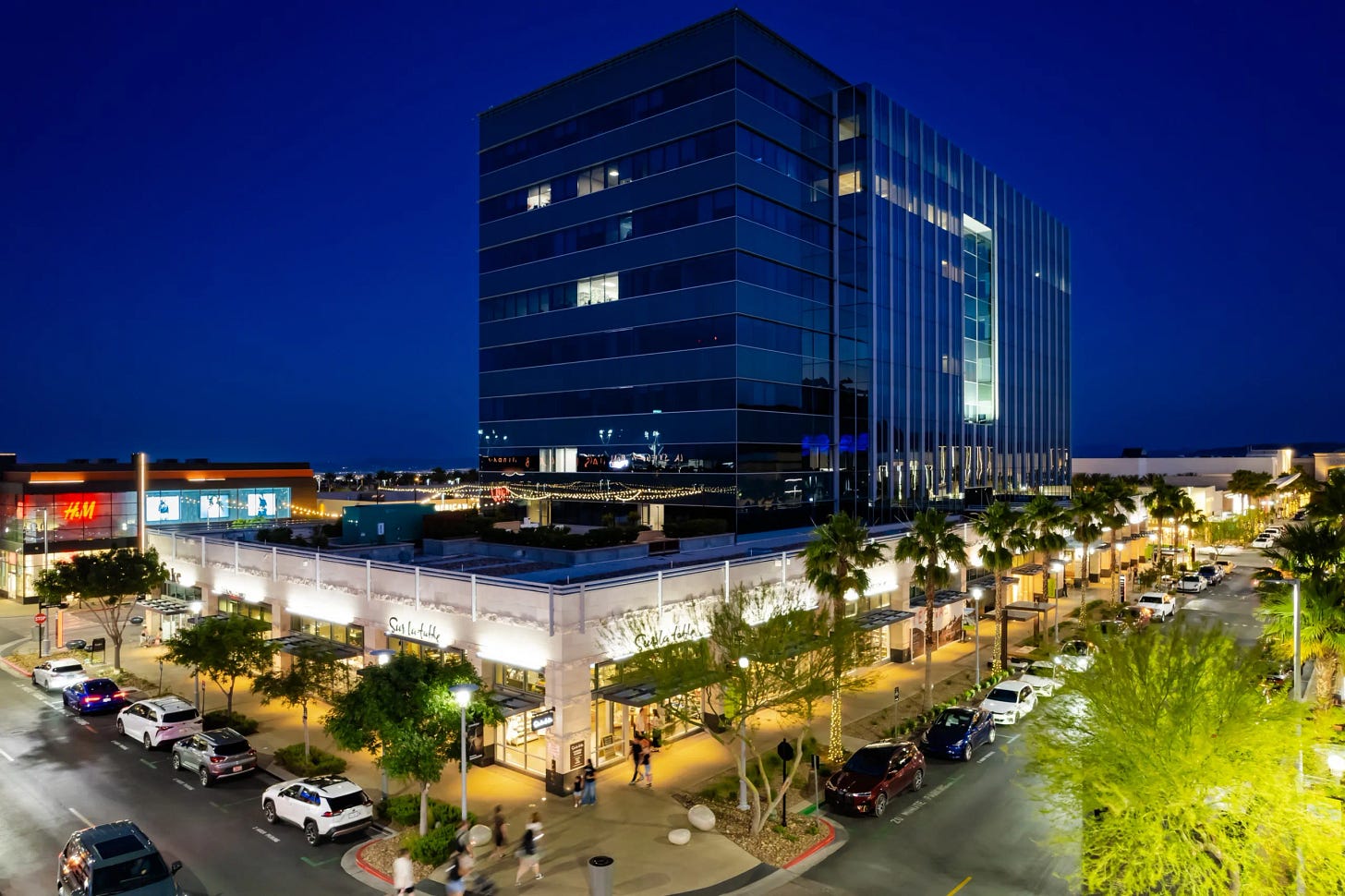 A modern glass office building rises behind a brightly lit shopping and dining area at night. Palm trees line the street, and cars are parked along both sides as people walk along the sidewalks.