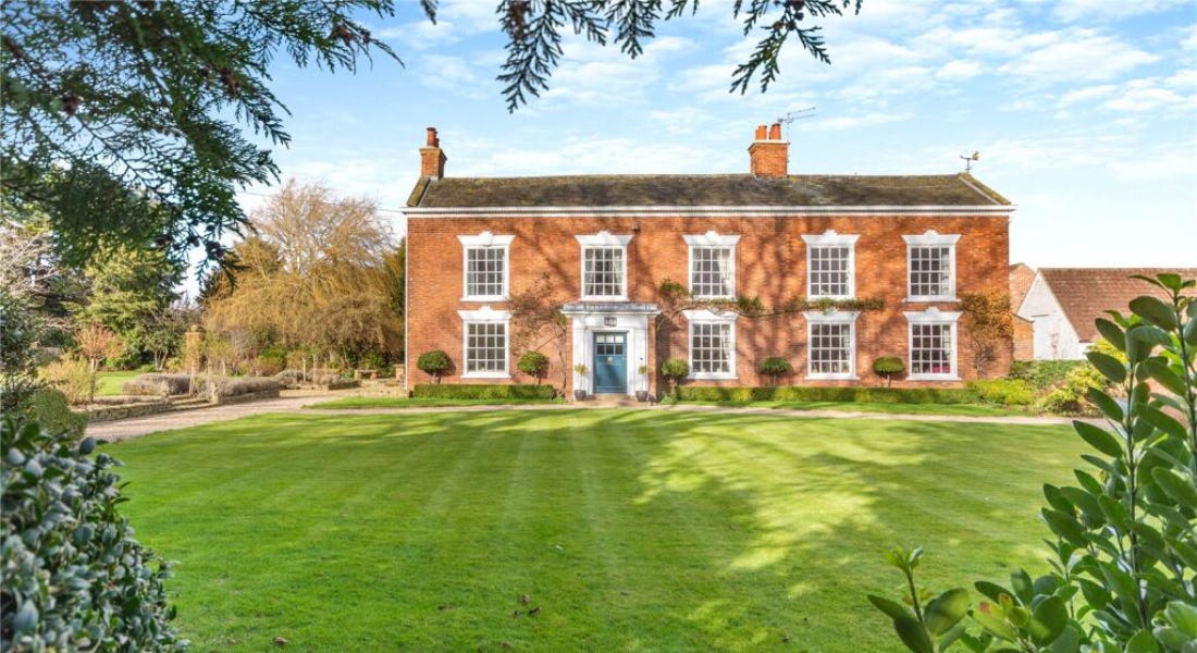 A large brick house with white windows, a sprawling green lawn, and trees and hedges
