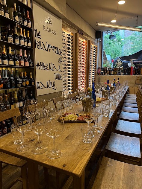 Photos inside In Vino wine bar in Yerevan, including a hand holding a bottle of Trinity Canyon Areni Noir, staff pouring wine behind shelves stocked with Armenian and international bottles, friends toasting with glasses of white wine, a long tasting table set with glassware beneath a sign reading 'The Renaissance of Armenian Wine Making,' and a wall of photos honoring winemakers