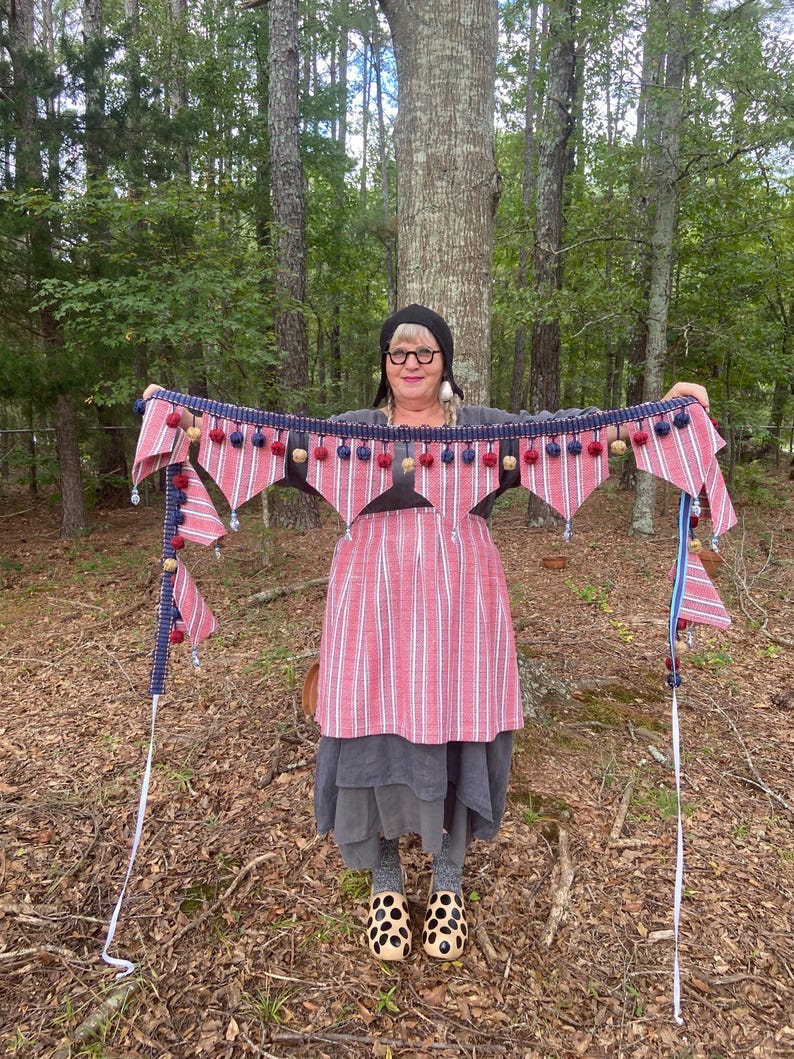 May include: A red and white striped banner with blue trim and decorative pom-poms. The banner has triangular pennants and is held up in front of a forest background. The banner is a decorative item. May include: A red and white striped banner with blue trim and decorative pom-poms. The banner has triangular pennants and is held up in front of a forest background. The banner is a decorative item.