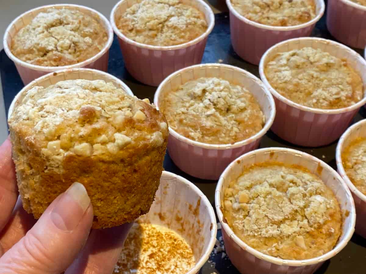 A hand holding an Apple, Oat and Cinnamon Muffin, with more muffins in the background. A hand holding an Apple, Oat and Cinnamon Muffin, with more muffins in the background.