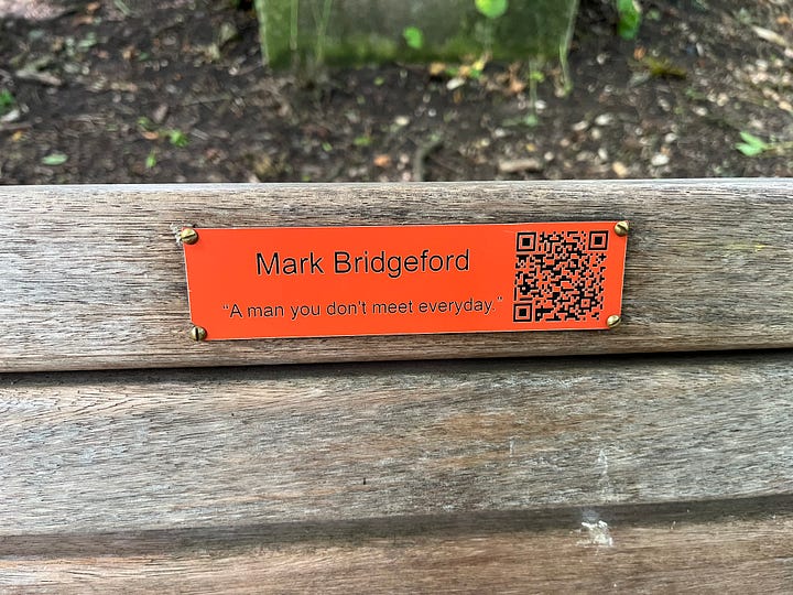 A bench with a bright orange label reading “Mark Bridgeford - A man you don’t meet every day.” with a QR code