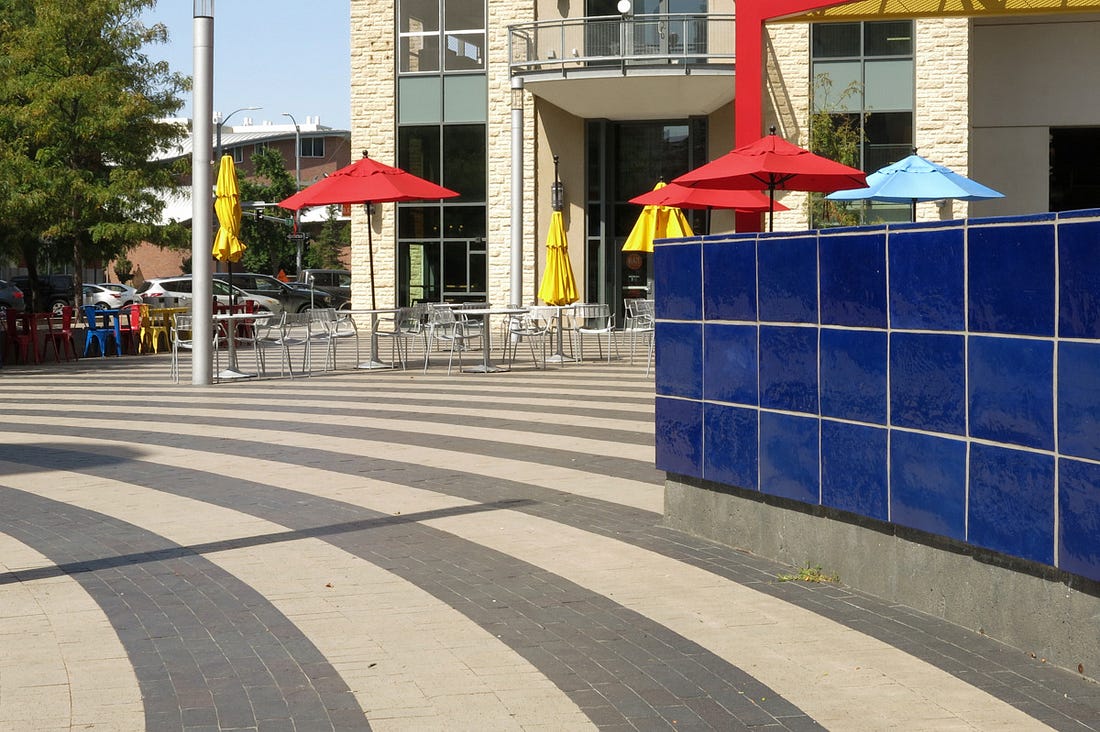 A photograph of a city square with a blue wall on the right, red and blue umbrellas over picnic tables, and paving stones set into dark and light stripes