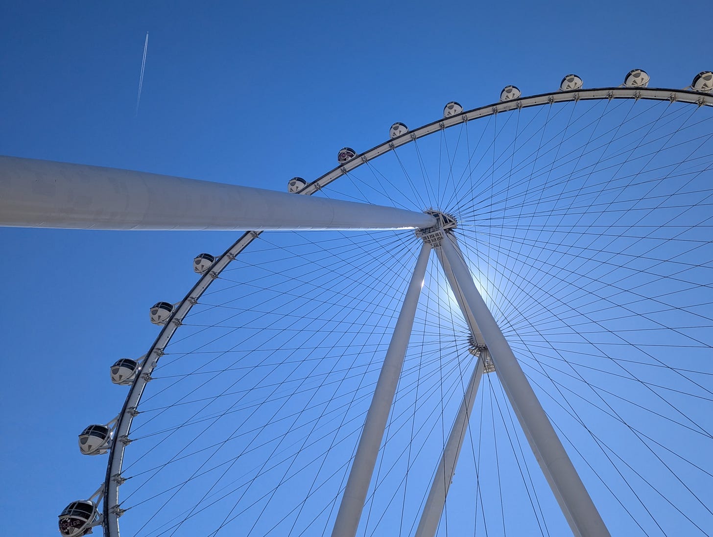 A photo of a "eye" type ferris wheel from below with the spokes making a cool pattern, especially with the sun behind the center beam. A jet leaves a contrail in the sky.