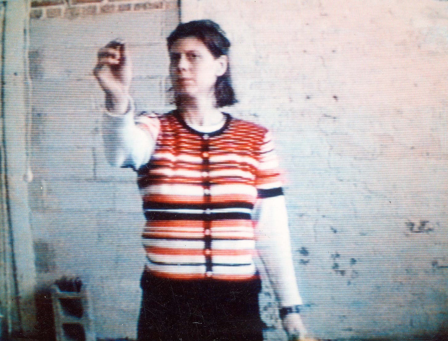 Lo-res video still of the artist, Mary Addison Hackett standing in an empty room facing the camera. Her right hand is extended as she prepares to throw a dart. She’s wearing a pink, white, and black striped shirt and black pants. Lo-res video still of the artist, Mary Addison Hackett standing in an empty room facing the camera. Her right hand is extended as she prepares to throw a dart. She’s wearing a pink, white, and black striped shirt and black pants.