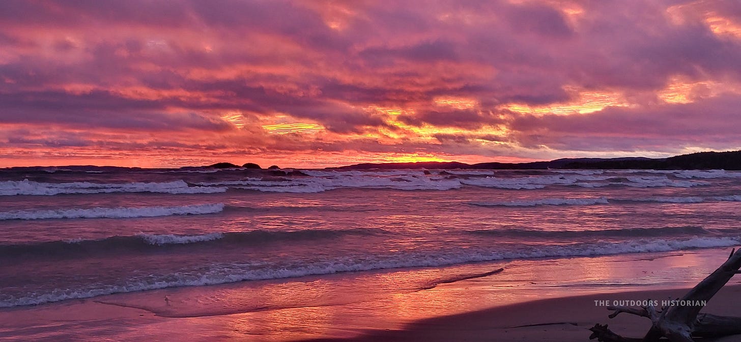 A vibrant sunset over the water. The sky is a wash of cotton-candy pink, orange, and purple clouds, which are perfectly reflected in the wet sand and calm water of the beach.