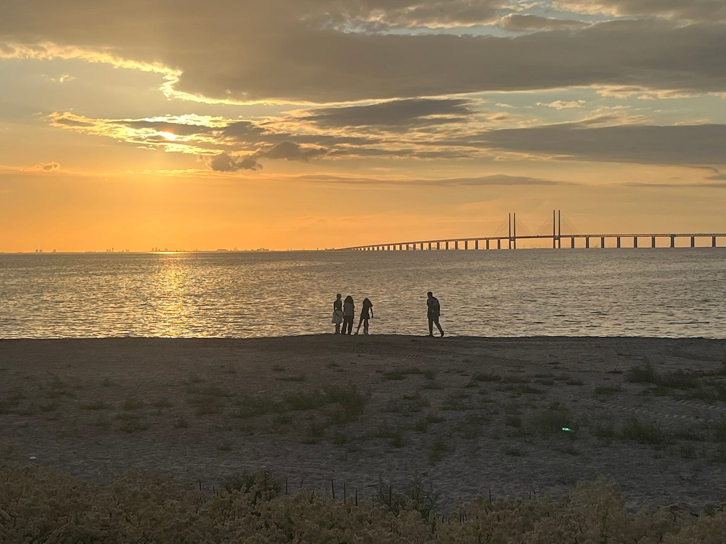 A beach in the late afternoon. The sky is partly clear and orange-ish but clouds cover the sun. There are 4 people on the beach in silhouette. There is a long bridge over the sea, covering half of the width of the picture, mostly low spans but with 2 tall spans. To the left, there are buildings, faintly visible across the sea.