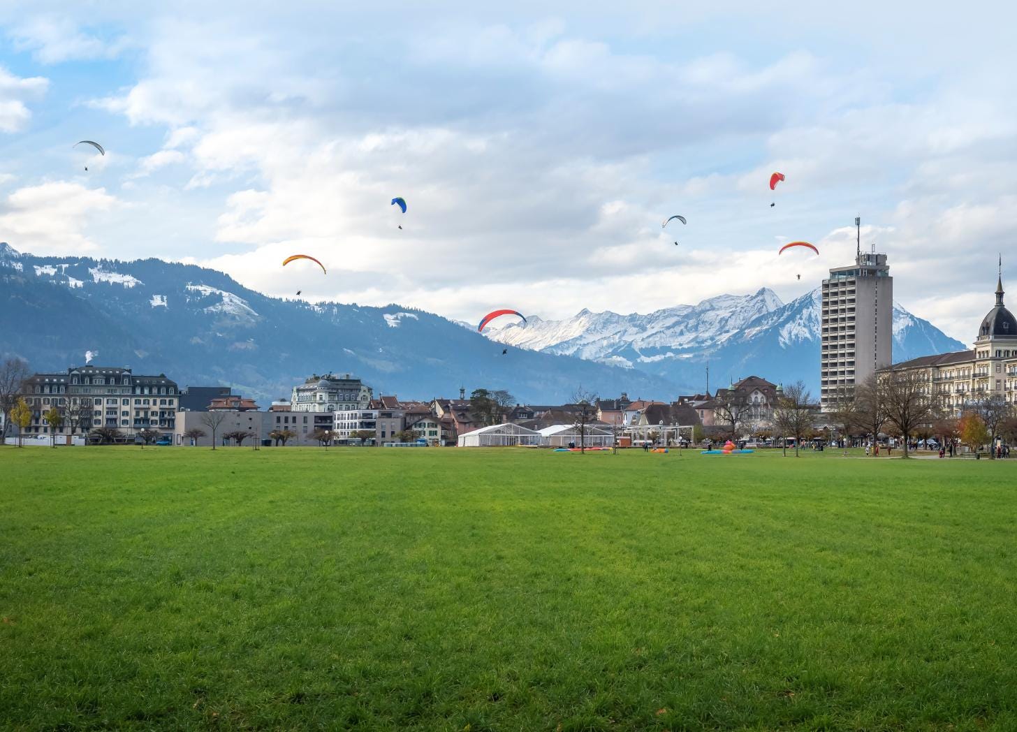 Wide view of the Höhematte Meadow in Interlaken with five colored paragliders descending in front of snow-capped Bernese Oberland mountains and the town's historic buildings. Wide view of the Höhematte Meadow in Interlaken with five colored paragliders descending in front of snow-capped Bernese Oberland mountains and the town's historic buildings.