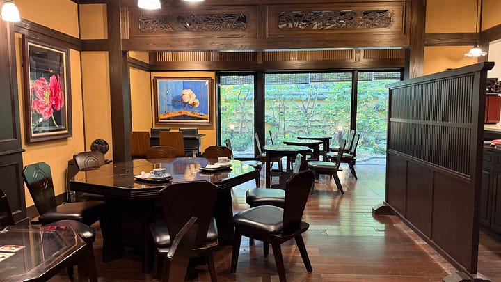 Image 1 alt text: A traditional Japanese dining room with dark wood tables and chairs, framed artwork on the walls, and large windows opening onto a leafy garden courtyard.  Image 2 alt text: A scenic shrine garden with a curved red bridge crossing a reflective pond, surrounded by trees and traditional wooden buildings.  Image 3 alt text: An accessible bathroom with a roll-in shower, bathtub, grab bars, handheld shower, raised toilet, sink with clearance underneath, and a blue shower chair.  Image 4 alt text: A modern hotel room with two twin beds, soft lighting, floor-to-ceiling curtains, and a small sitting area with a round table and armchair.