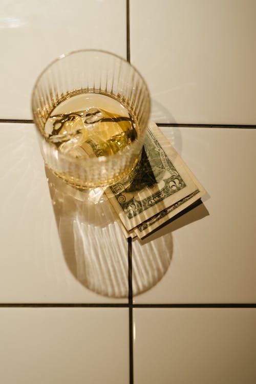 Free A close-up of a glass of whiskey with ice on top of dollar bills, casting shadows on white tiles. Stock Photo