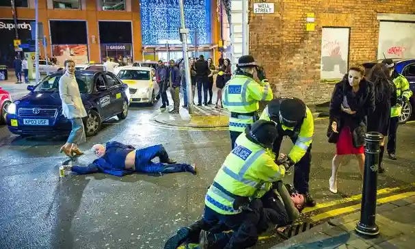 A crowded street with several police officers dealing with a drunk man down on the ground, another man fighting an arrest, strangers stop to look at the chaos in the middle of the busy road in Manchester, England.