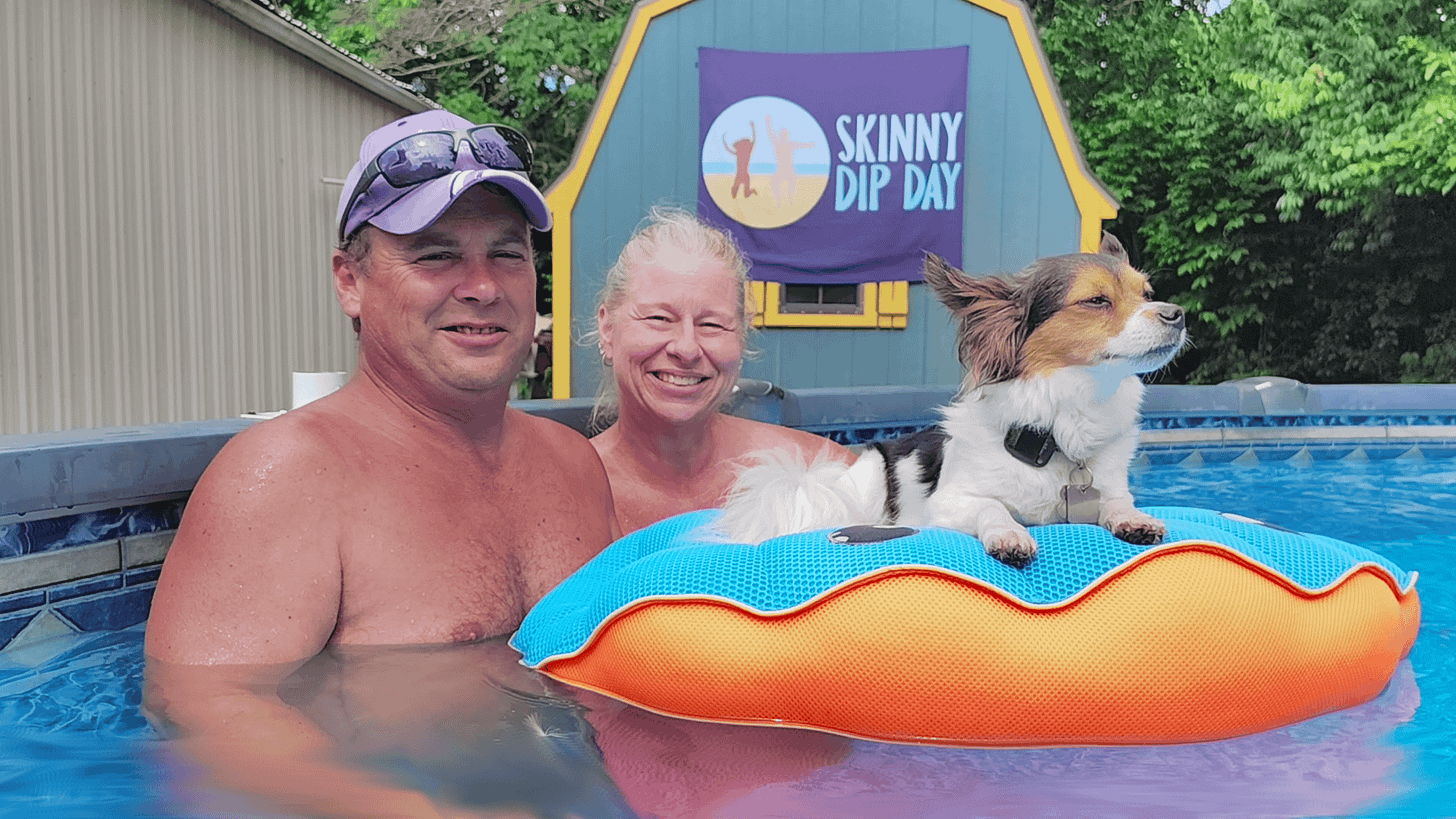 Alt text: A smiling couple relaxes in an above-ground pool, the man in a purple ball cap with sunglasses propped on the brim and a woman with long blonde hair beside him. A small white, black, and tan dog perches on an orange-and-blue pool float in the foreground. Behind them, a purple Skinny Dip Day banner hangs on a blue and yellow shed. Alt text: A smiling couple relaxes in an above-ground pool, the man in a purple ball cap with sunglasses propped on the brim and a woman with long blonde hair beside him. A small white, black, and tan dog perches on an orange-and-blue pool float in the foreground. Behind them, a purple Skinny Dip Day banner hangs on a blue and yellow shed.