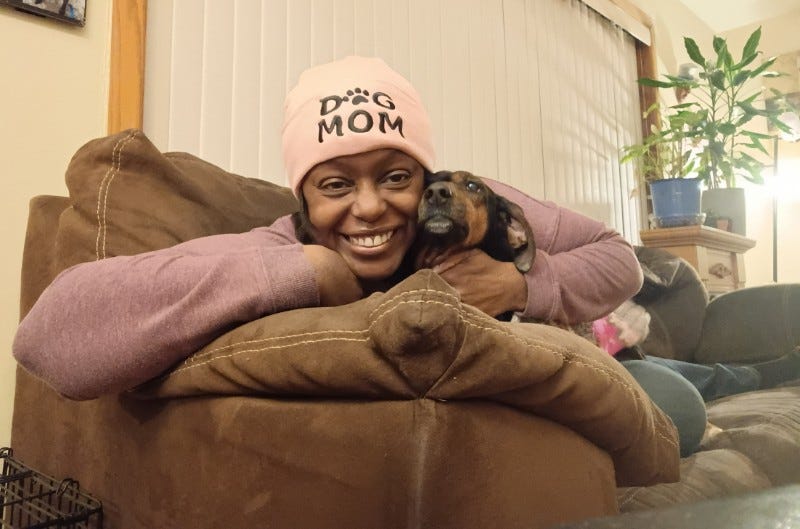 Shamontiel wears a "dog mom" pink hat while holding onto her Dachshund/Hound mix on a couch.