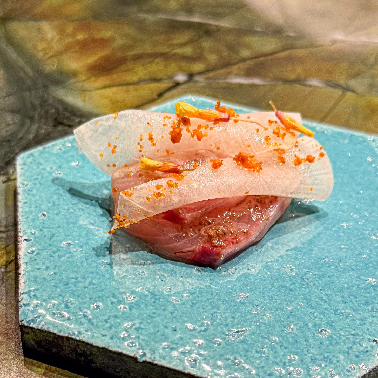 Sliced raw amberjack sashimi with orange roe garnish and edible flowers on turquoise ceramic plate at Mantra restaurant