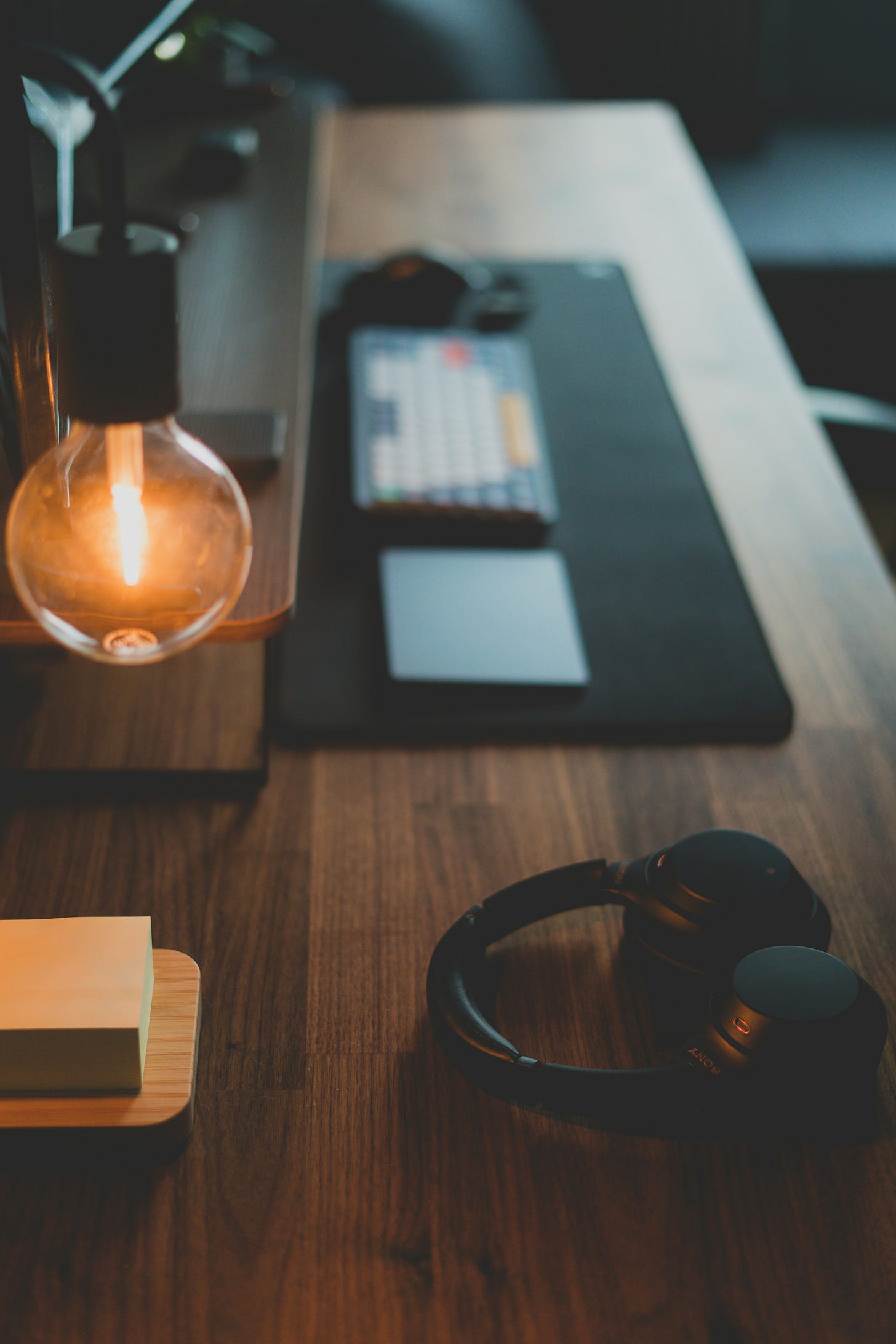 A warm, softly lit workspace featuring a glowing exposed lightbulb, a pair of black headphones resting on a wooden desk, and a blurred keyboard and trackpad in the background. The scene suggests quiet focus and creative work. A warm, softly lit workspace featuring a glowing exposed lightbulb, a pair of black headphones resting on a wooden desk, and a blurred keyboard and trackpad in the background. The scene suggests quiet focus and creative work.