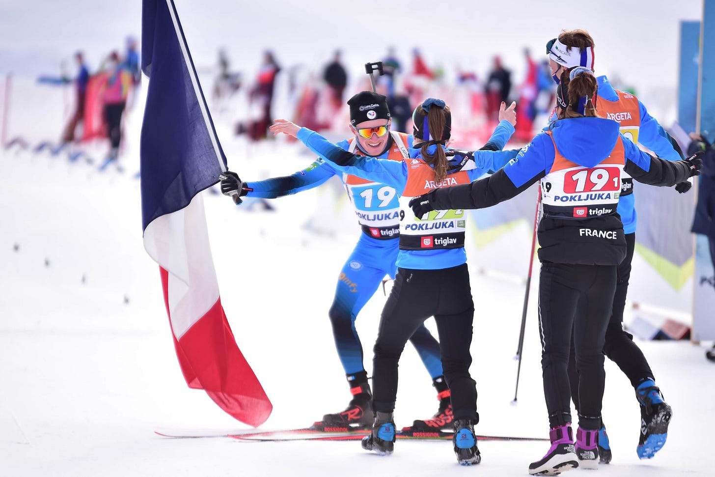 French biathlon relay team celebrating at the finish line in Pokljuka, with athletes in blue reaching out for hugs.