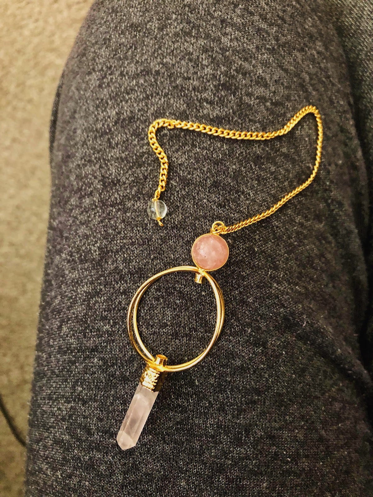 A pink quartz pendulum on a gold chain sitting on a sweatpants'd lap